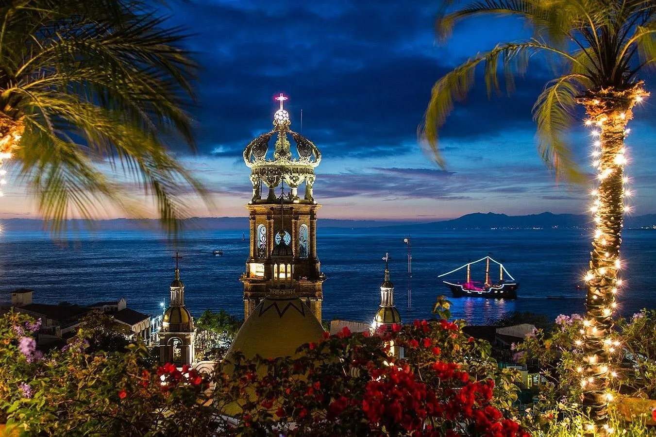 The crown of the Guadalupe church in Puerto Vallarta at dusk, with the lit-up pirate ship on the bay behind