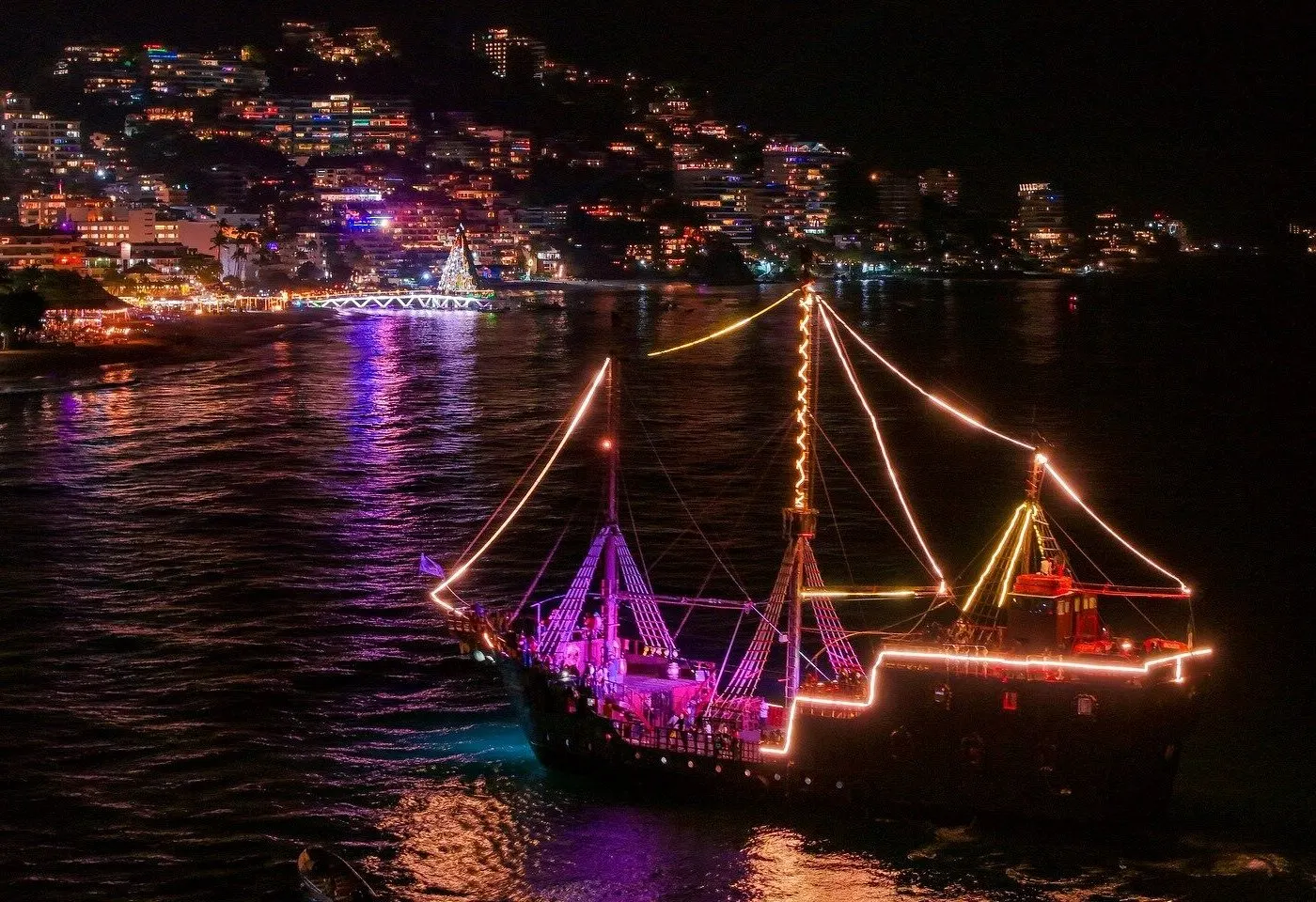 The pirate ship outlined in lights at night, with the Los Muertos pier and hillside lights of Puerto Vallarta behind
