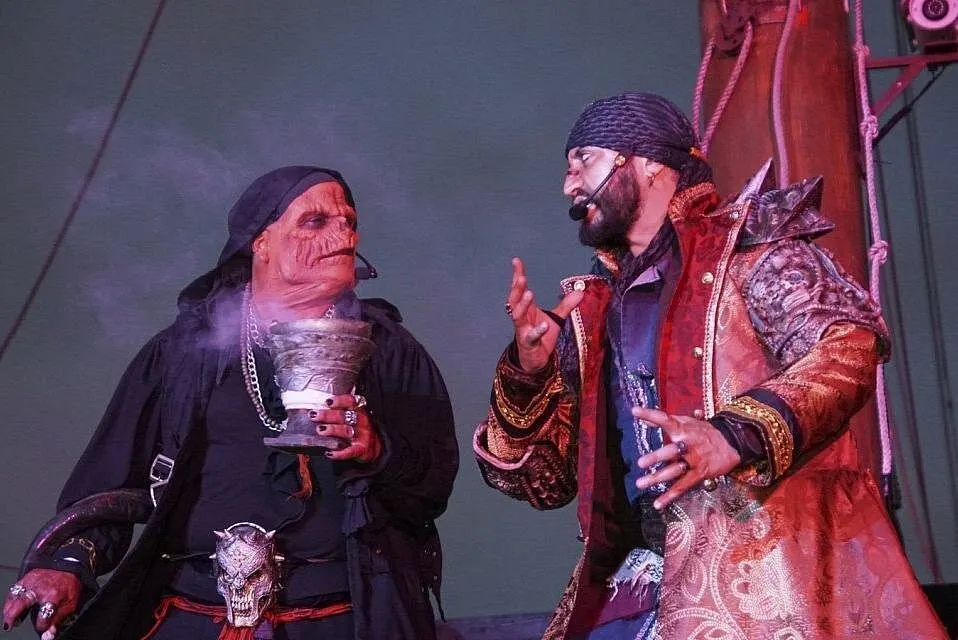 The pirate captain in a gold brocade coat faces a masked pirate holding a smoking goblet in the night show